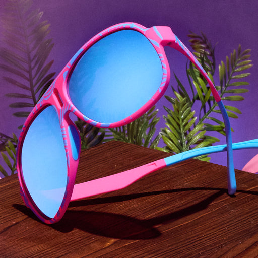 A pair of pink and blue tropical Super Fly shades is displayed on a wooden table, set against a backdrop of tropical greenery.