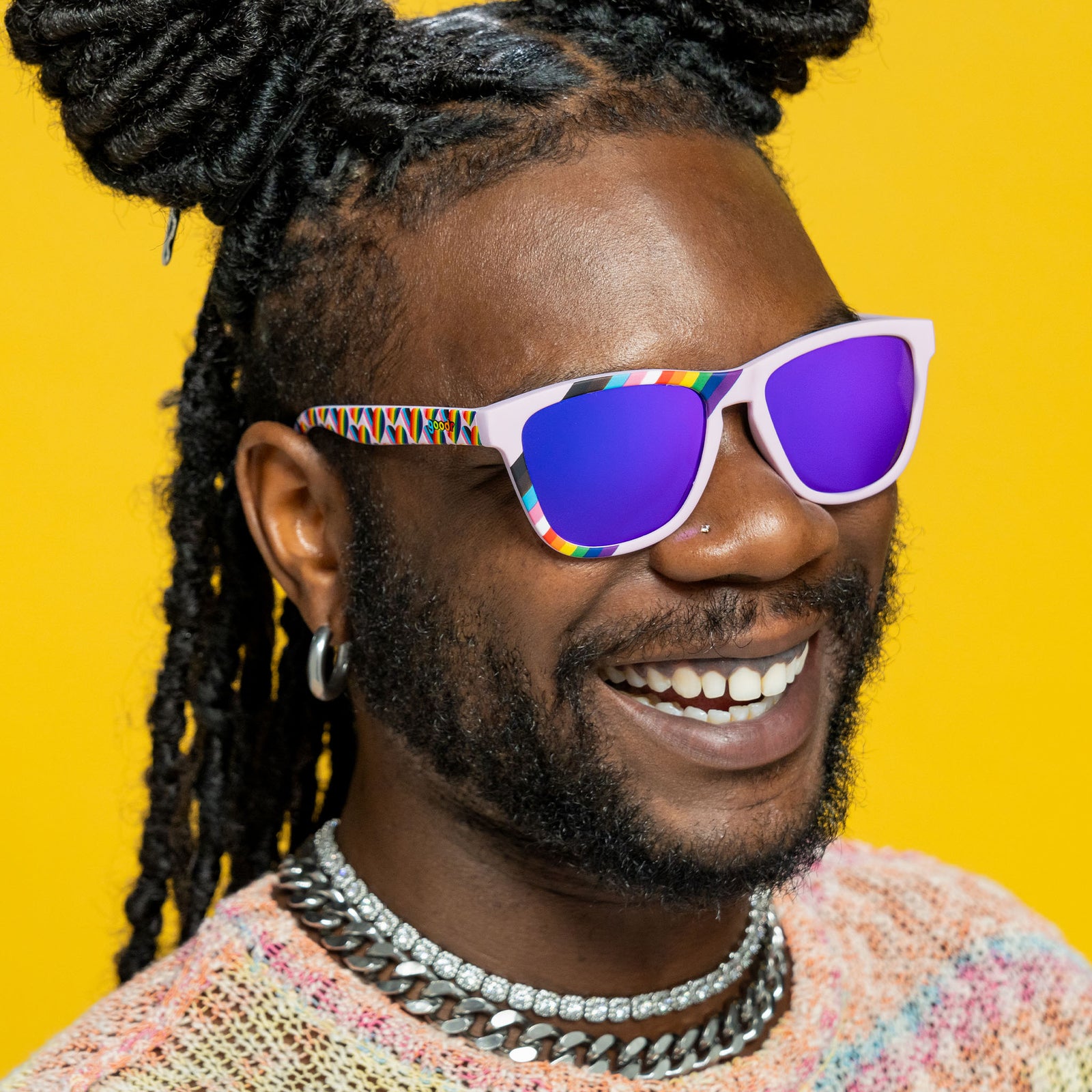 Three-quarter angle view of a happy young man wearing lilac rainbow flag LGBTQ+ Pride sunglasses with purple lenses.